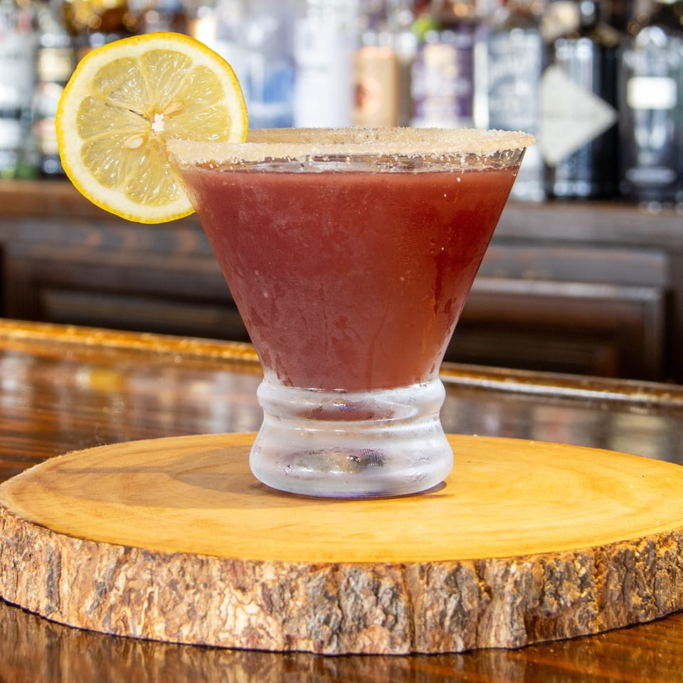 Red cocktail with a sugar-rimmed glass and lemon garnish on a wooden slab at a bar.