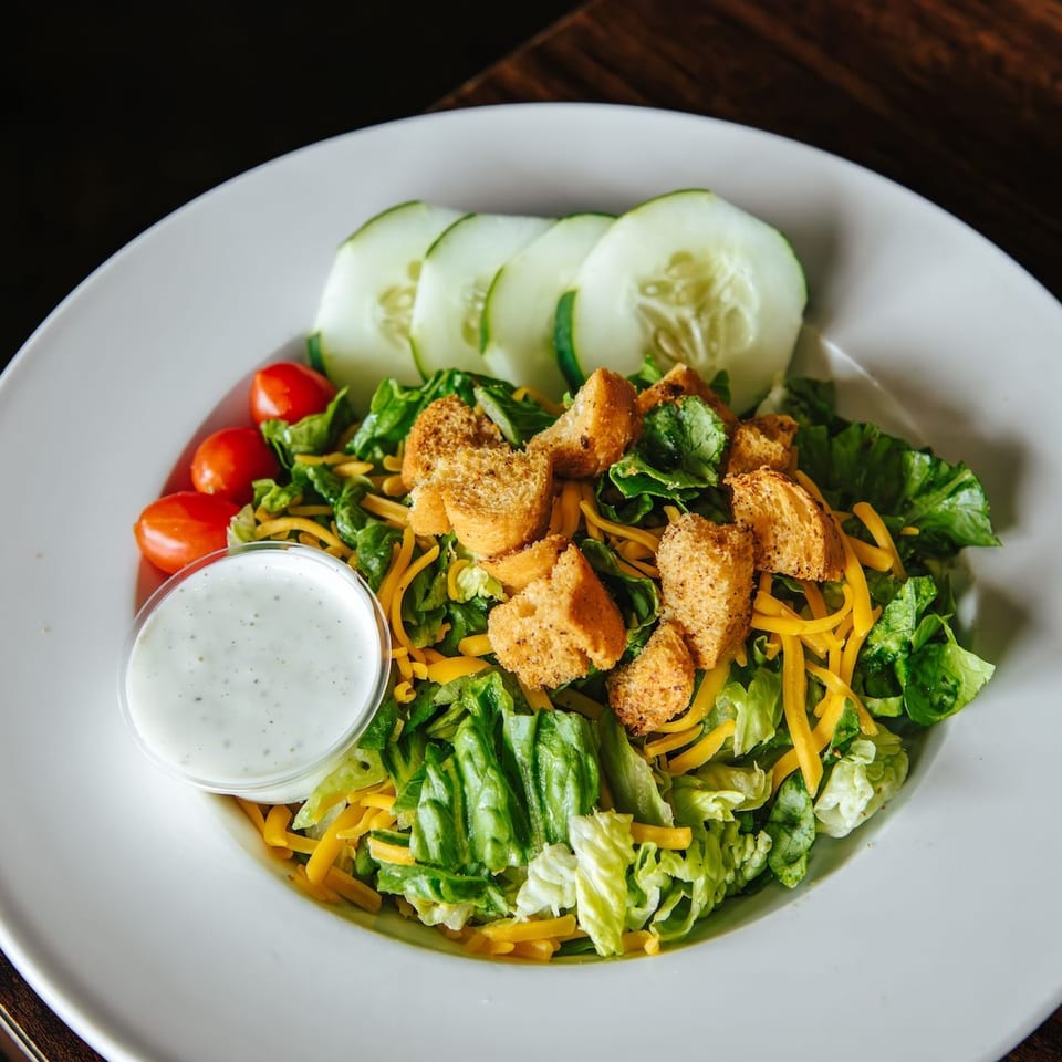 Fresh green salad with cucumber, cherry tomatoes, shredded cheese, croutons, and creamy dressing on a white plate.