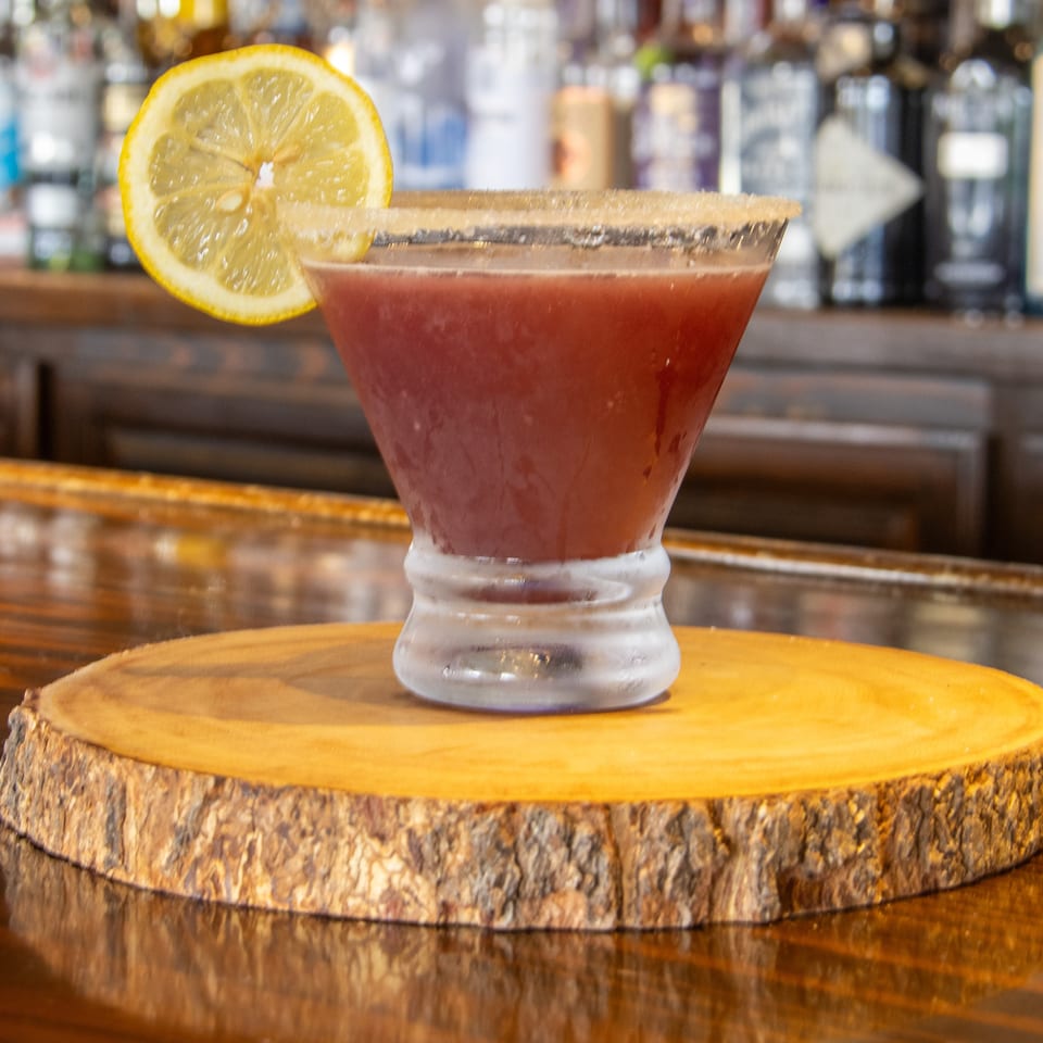 Red cocktail in a sugar-rimmed glass garnished with a lemon slice, displayed on a wooden coaster at a bar.