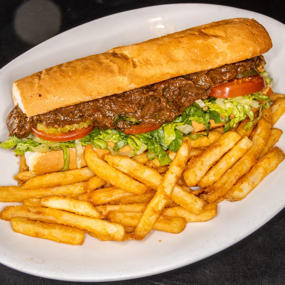 Steak sandwich with lettuce, tomatoes, and pickles served alongside golden French fries on a white plate.