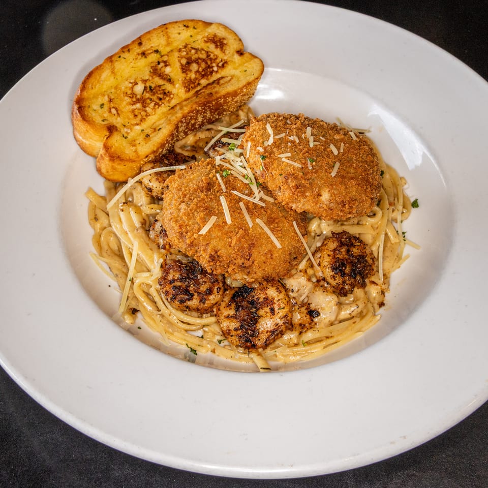 Breaded chicken and grilled shrimp over creamy pasta, topped with Parmesan, served with garlic toast on a white plate.