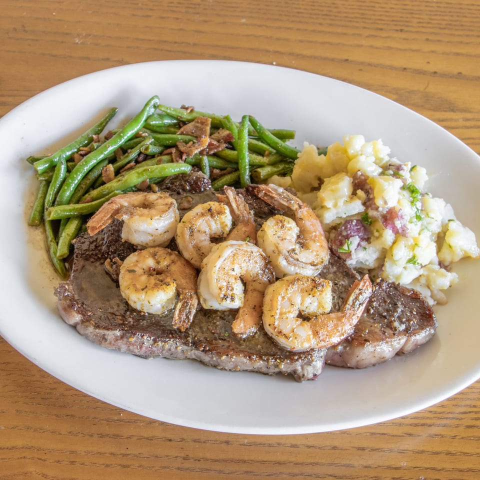 Grilled steak topped with shrimp, served with green beans and loaded mashed potatoes on a white plate.