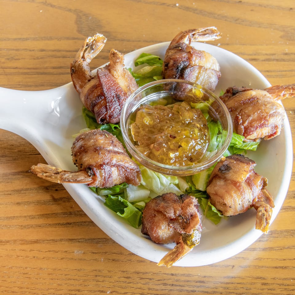 Bacon-wrapped shrimp arranged on lettuce around a glass bowl of dipping sauce on a white platter.