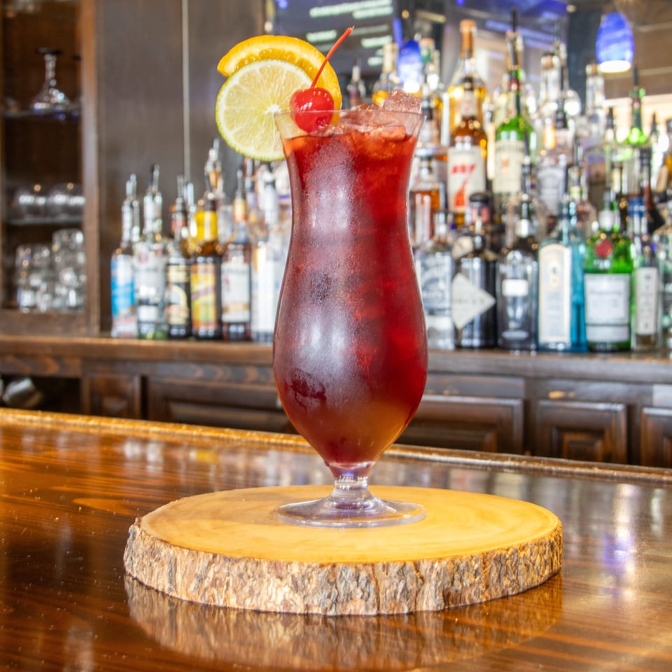 Red iced cocktail garnished with lemon slices and a cherry, displayed on a wooden coaster at a fully stocked bar.