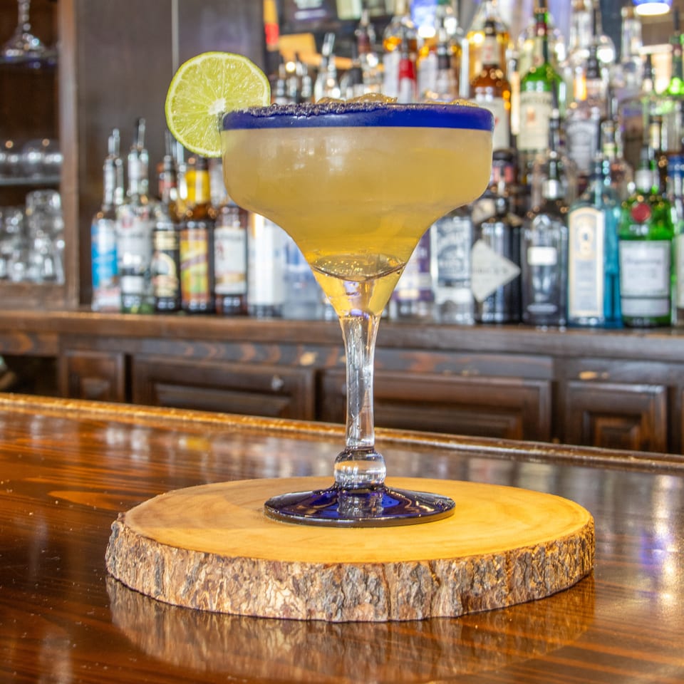 Margarita with a blue salt rim and lime garnish sits on a wooden coaster at a bar lined with liquor bottles.