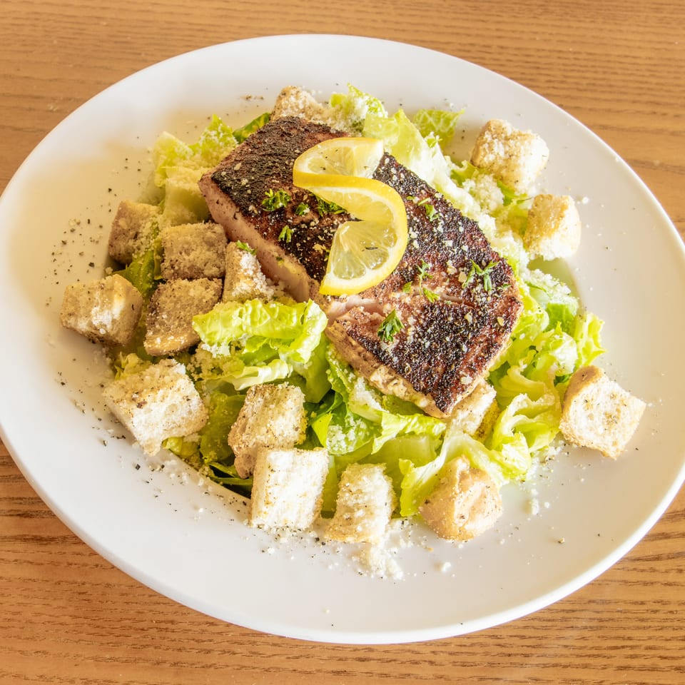 Grilled salmon fillet with lemon slices atop a Caesar salad with romaine lettuce, croutons, and Parmesan cheese.