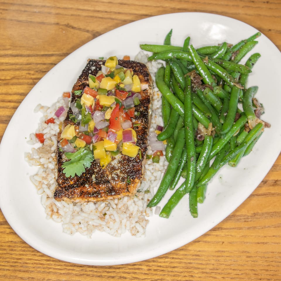 Blackened salmon topped with mango salsa, served over rice with seasoned green beans on a white plate.