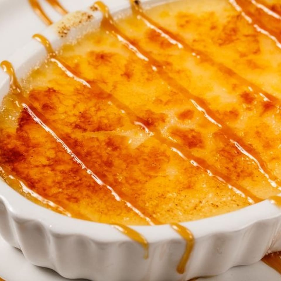 Caramelized crème brûlée in a white fluted ramekin, drizzled with caramel sauce.