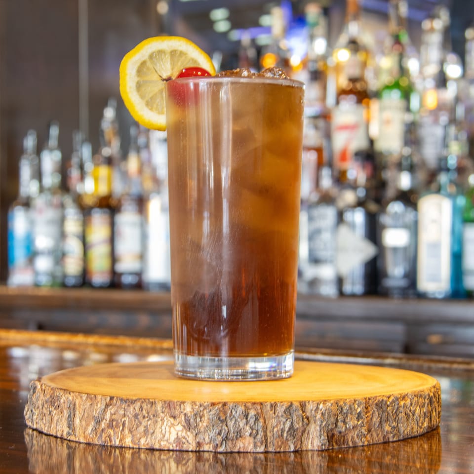 Tall amber cocktail garnished with a lemon wheel and cherry, displayed on a wooden slab before a warmly lit bar.