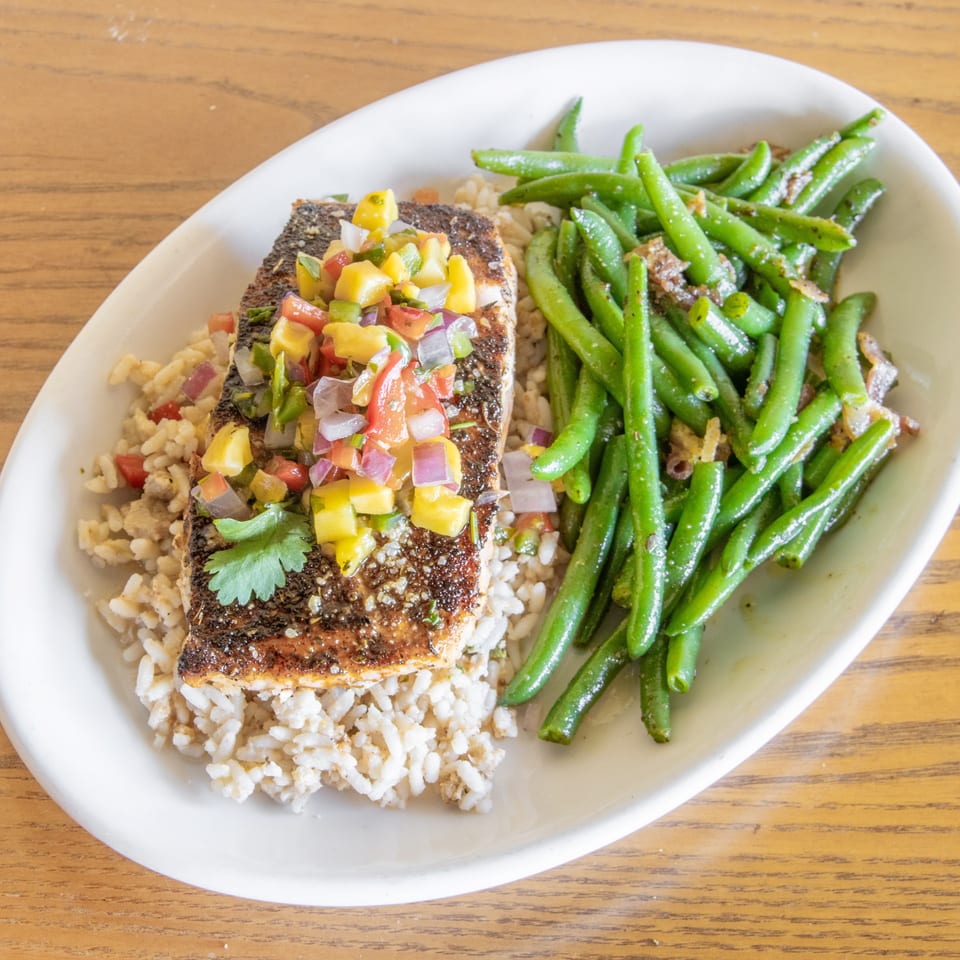 Seasoned grilled fish topped with colorful salsa, served over rice with sautéed green beans on a white plate.