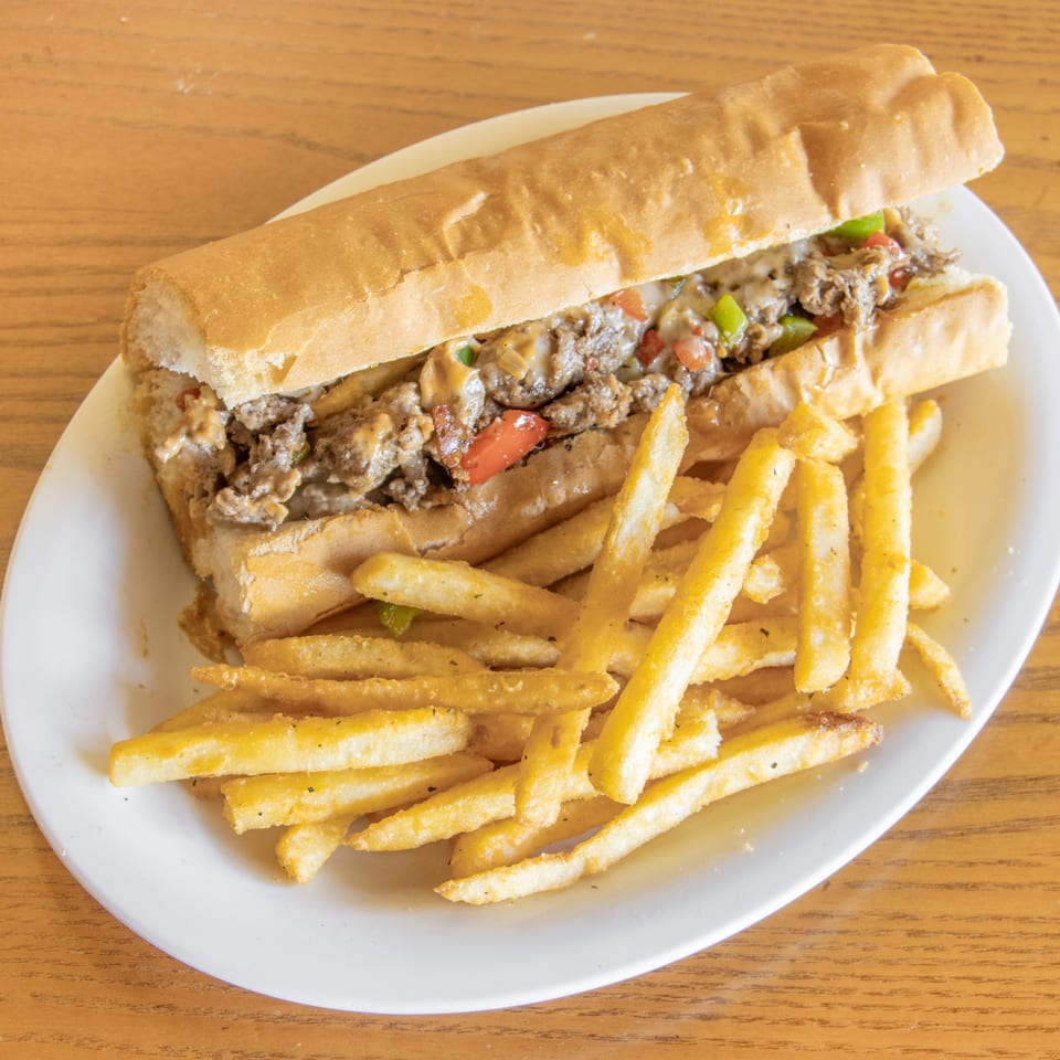 Loaded steak sandwich with sautéed vegetables and fries served on a white plate atop a wooden table.